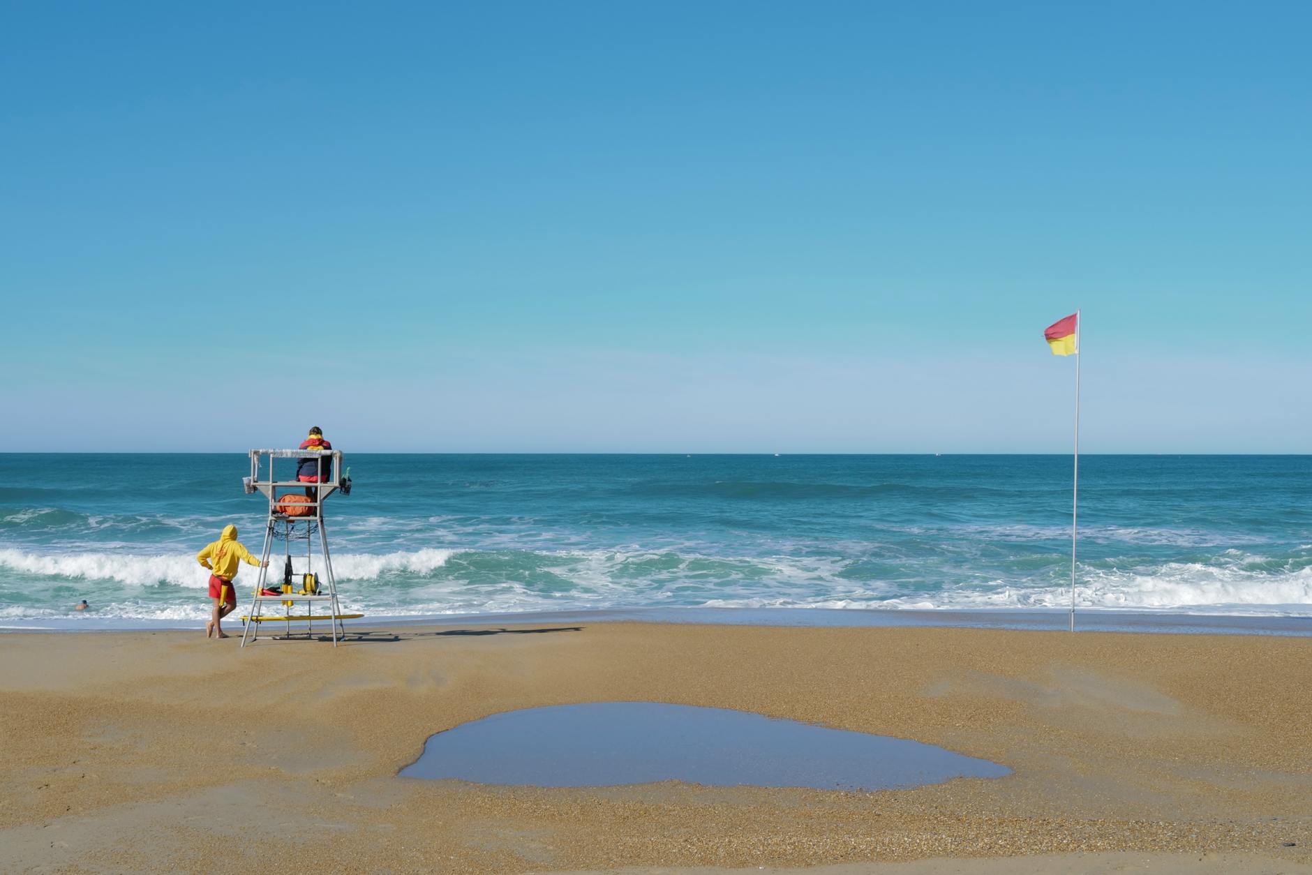 Serene beach scene with lifeguards, waves, and safety flag under a clear sky.