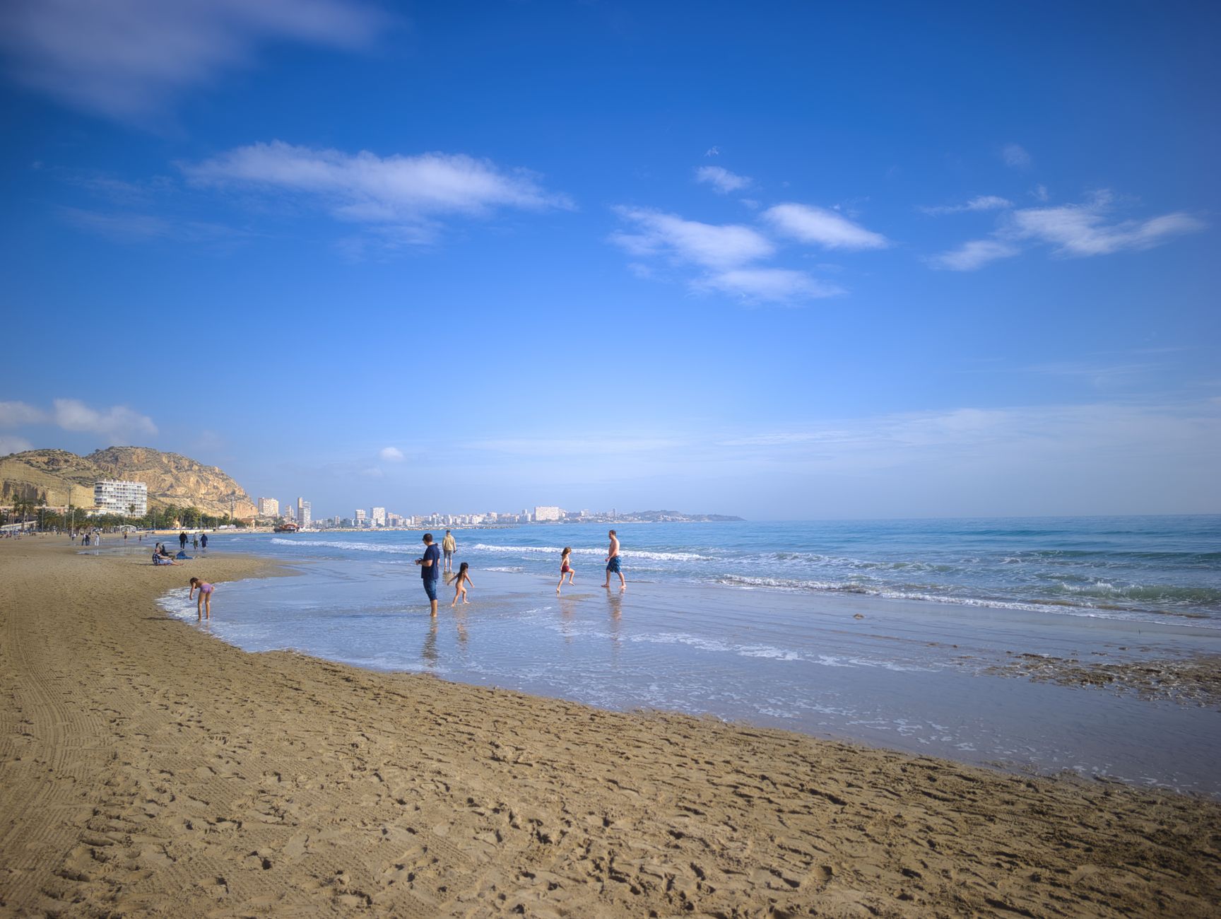 A scenic view of a family playing on the beach in Alicante, Spain under a clear blue sky.