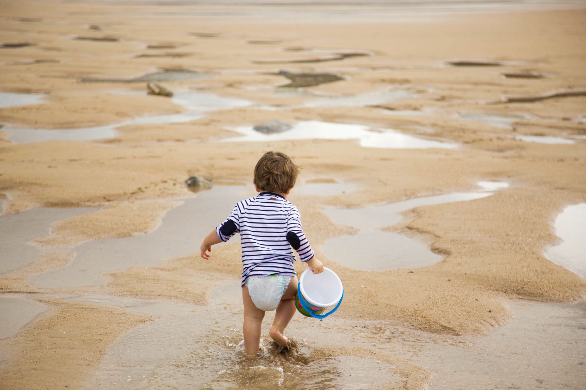 A child carrying a bucket walks along the sandy beach on a sunny day, enjoying leisure time.
