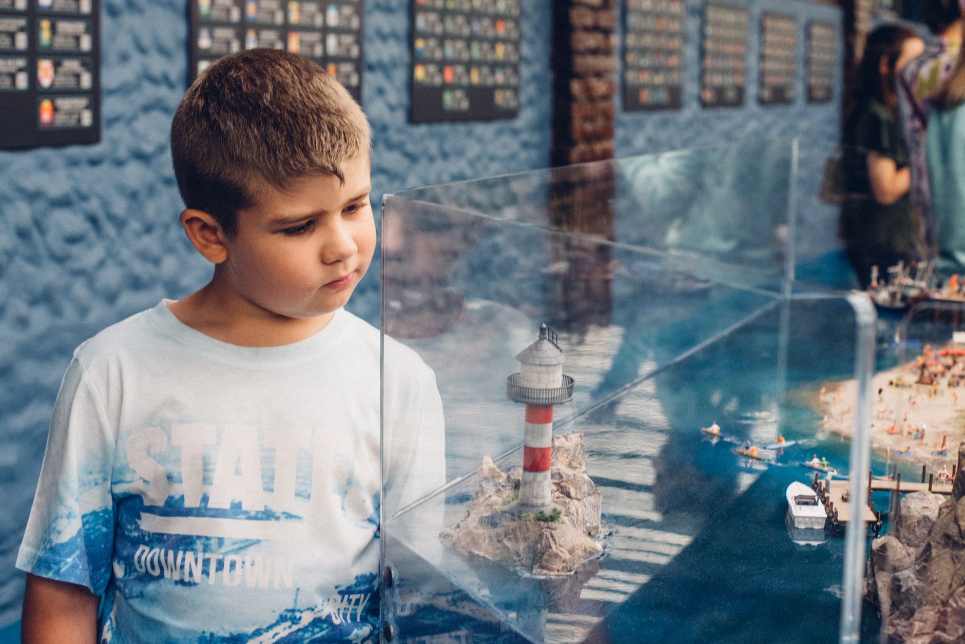 Curious child gazes at a detailed miniature lighthouse in a museum display.