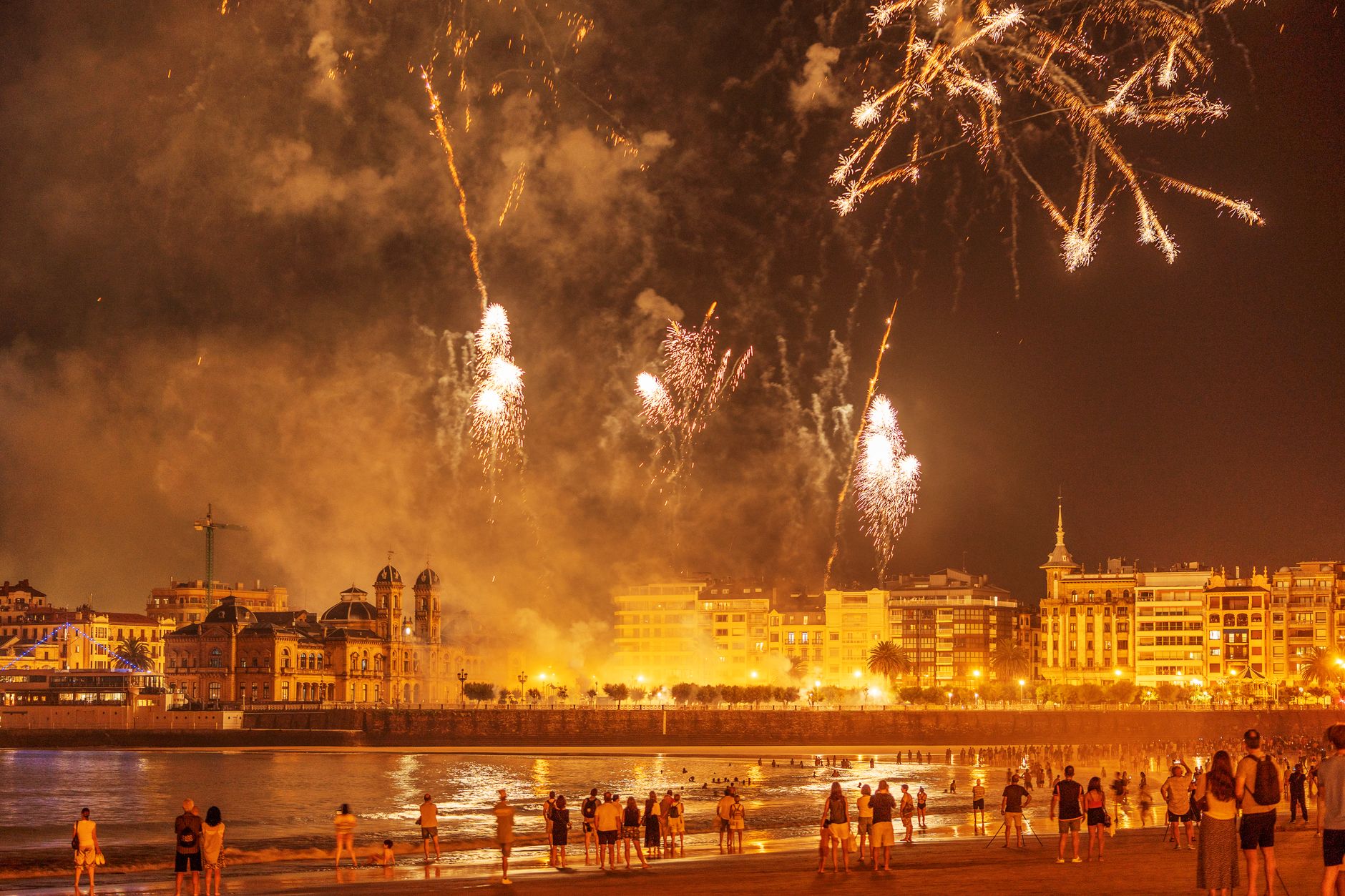 Crowds enjoy vibrant fireworks over the coastal city at night, creating a festive atmosphere.