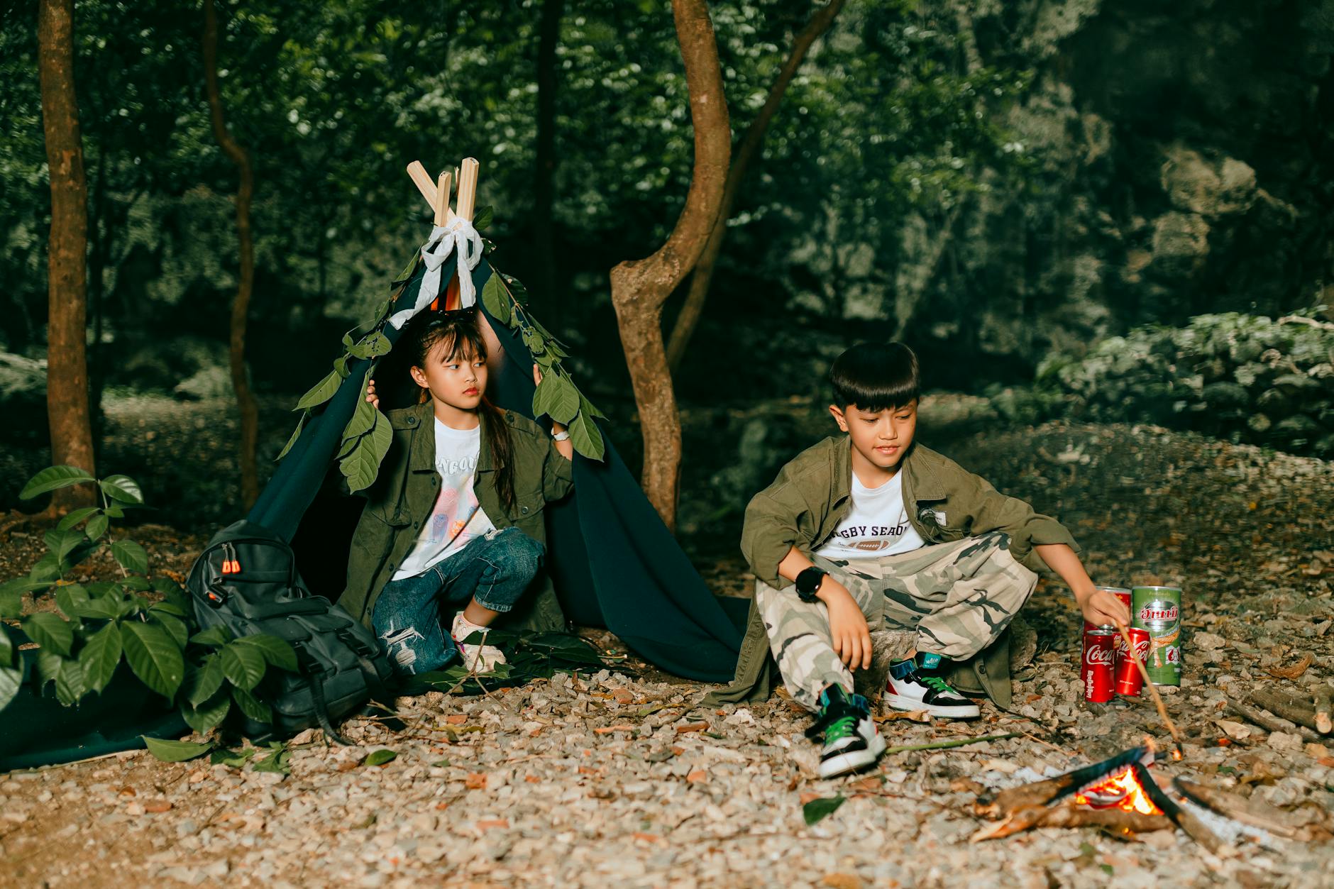 Two children relaxing by a campfire near a tent in a forest setting, enjoying nature.
