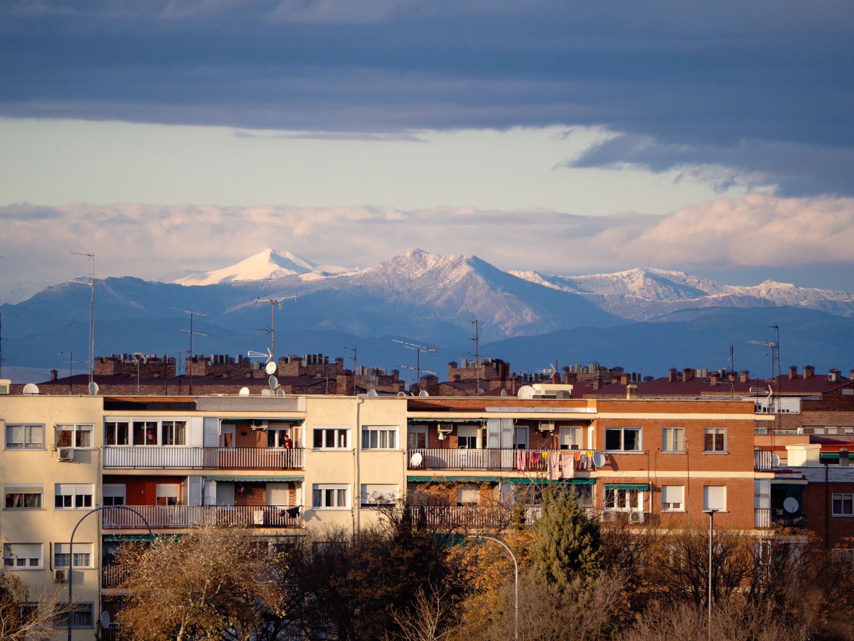 Urban view of Madrid with distant snow-capped mountains under a dramatic sky.