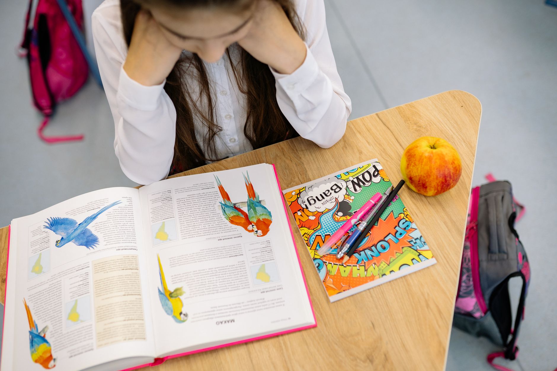 A young student studying at a desk with an open book, colorful notebook, pens, and an apple.