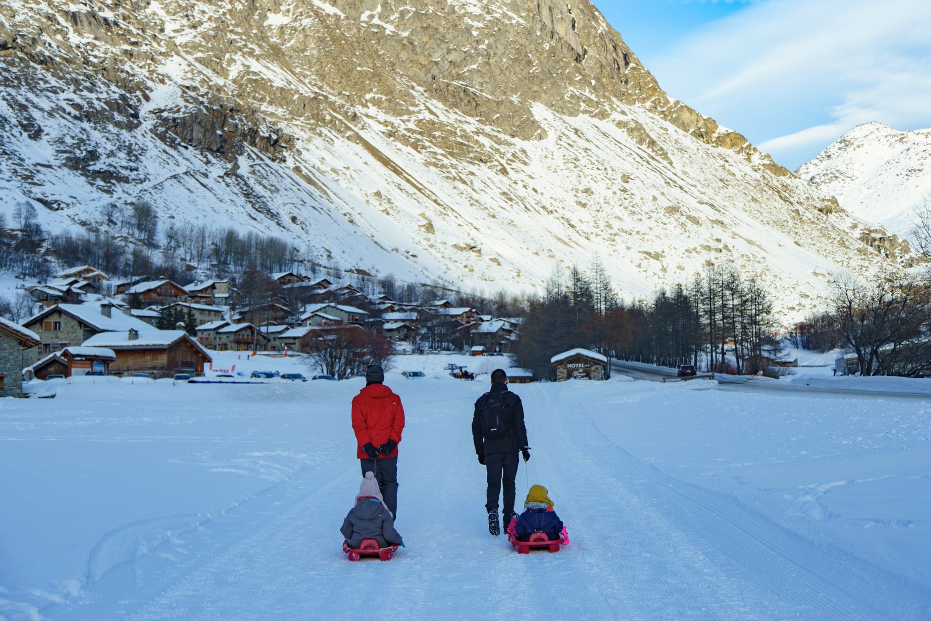 A family enjoys a winter day sledding along a snowy path in a picturesque village beneath snowy mountains.