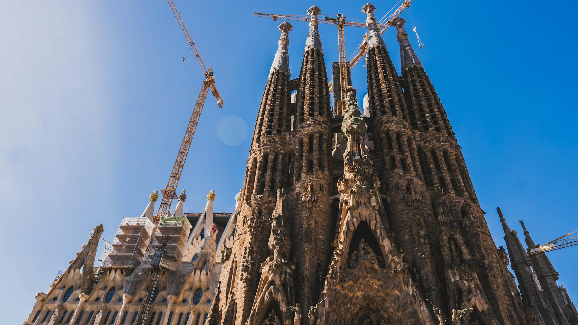 View of Sagrada Familia&rsquo;s intricate facade and cranes against a clear blue sky in Barcelona.