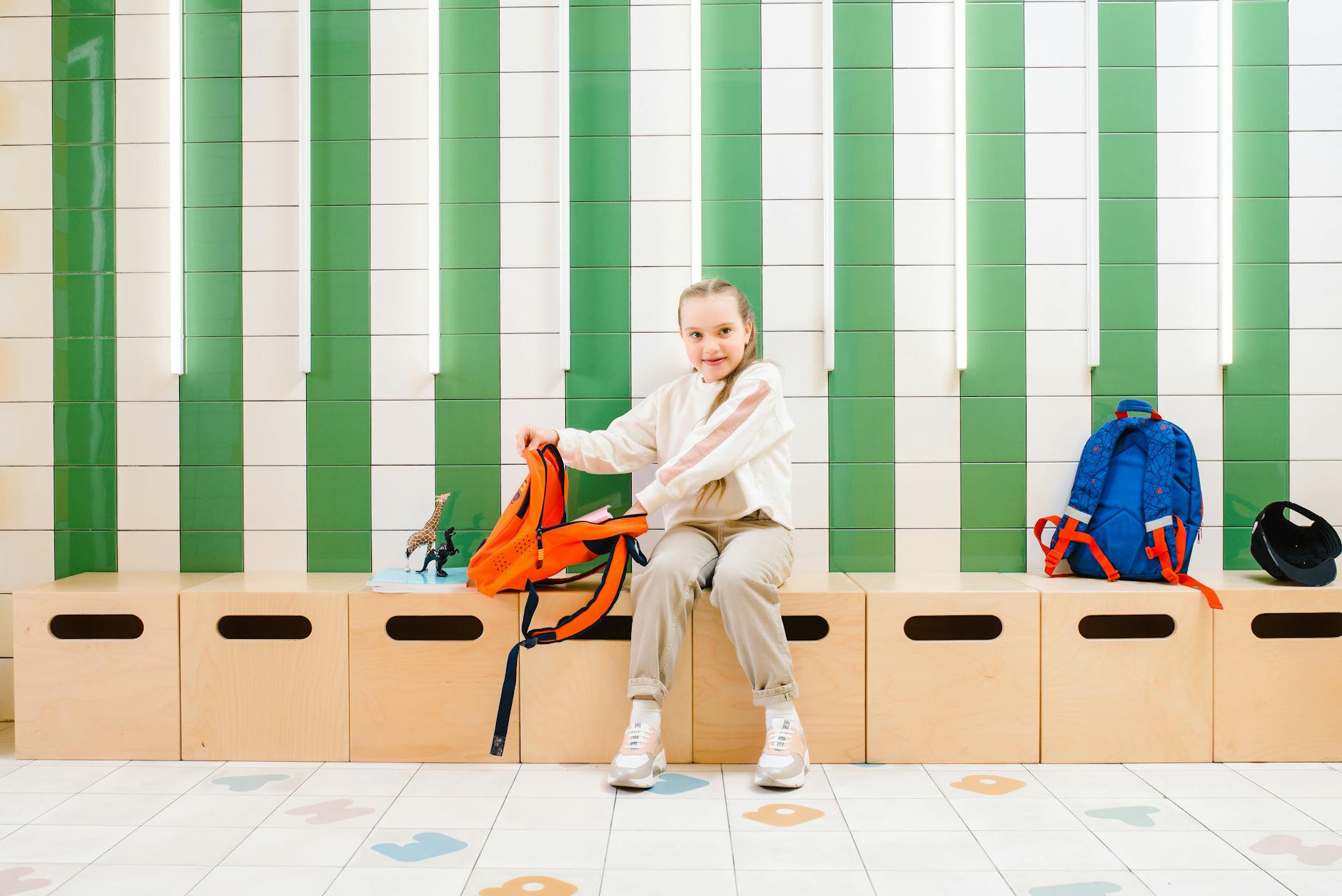 A young girl smiles while packing her backpack in a school locker room with bright decor.