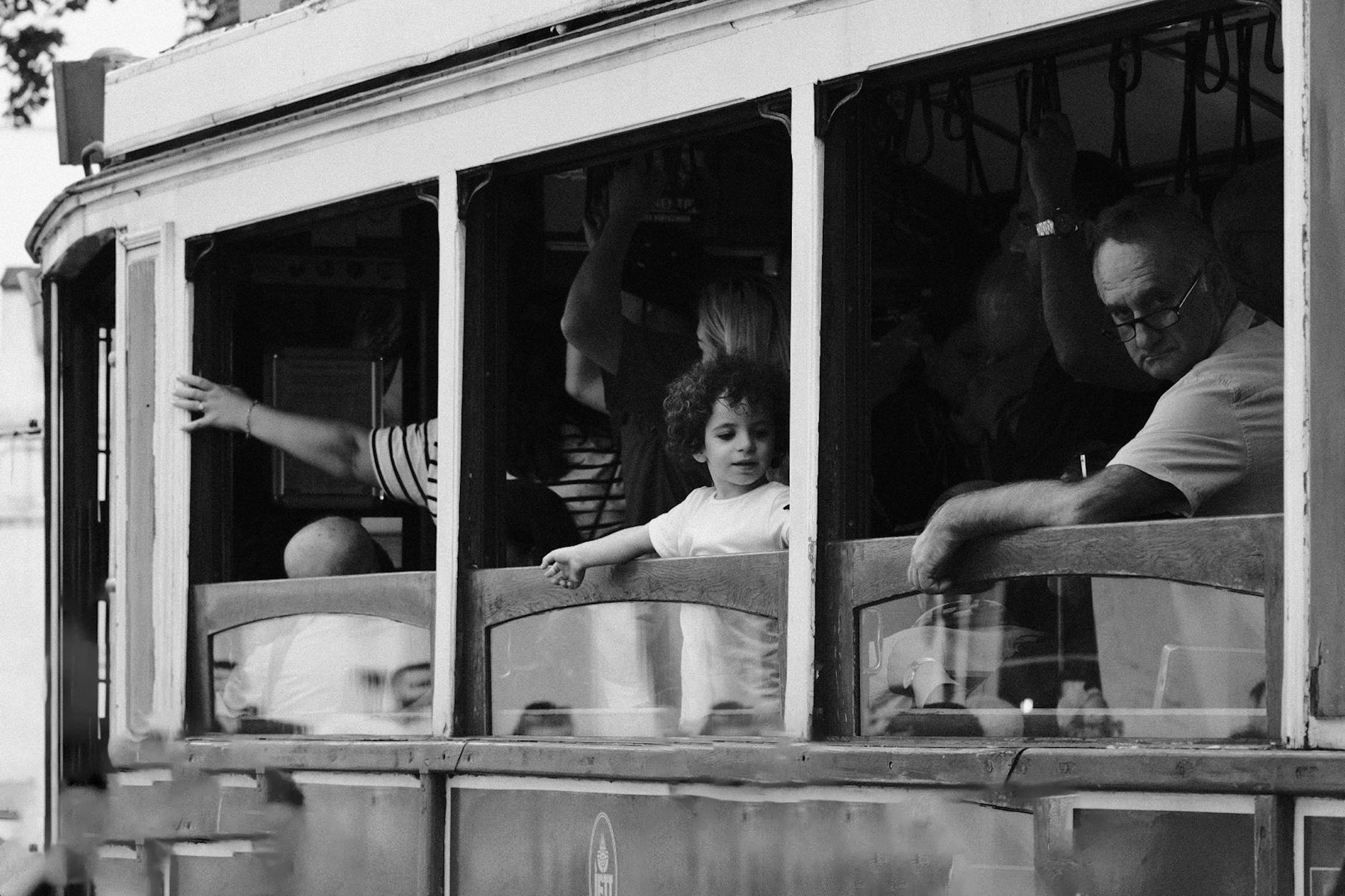 Black and white photo of a child gazing out of a tram window with adults around, showcasing a nostalgic travel moment.