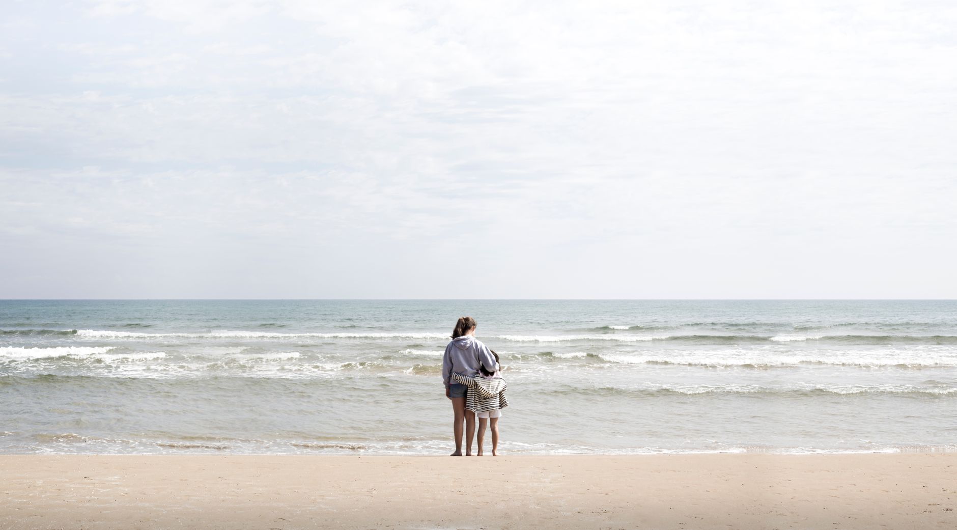 A mother and child stand at the shoreline of a Valencia beach, gazing at the waves.