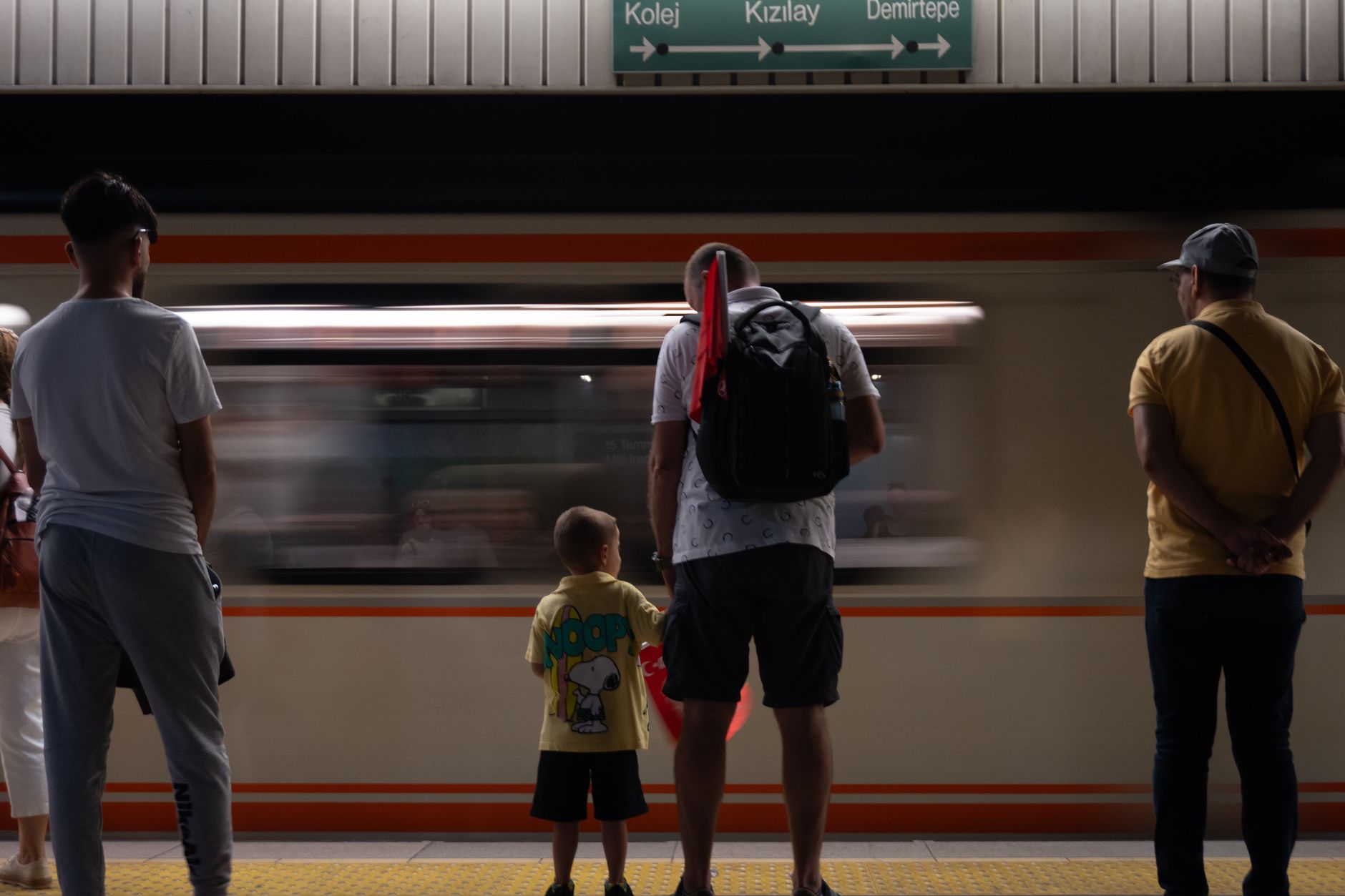 Father and son stand on Ankara metro platform, blurred train passing by, creating a sense of motion.