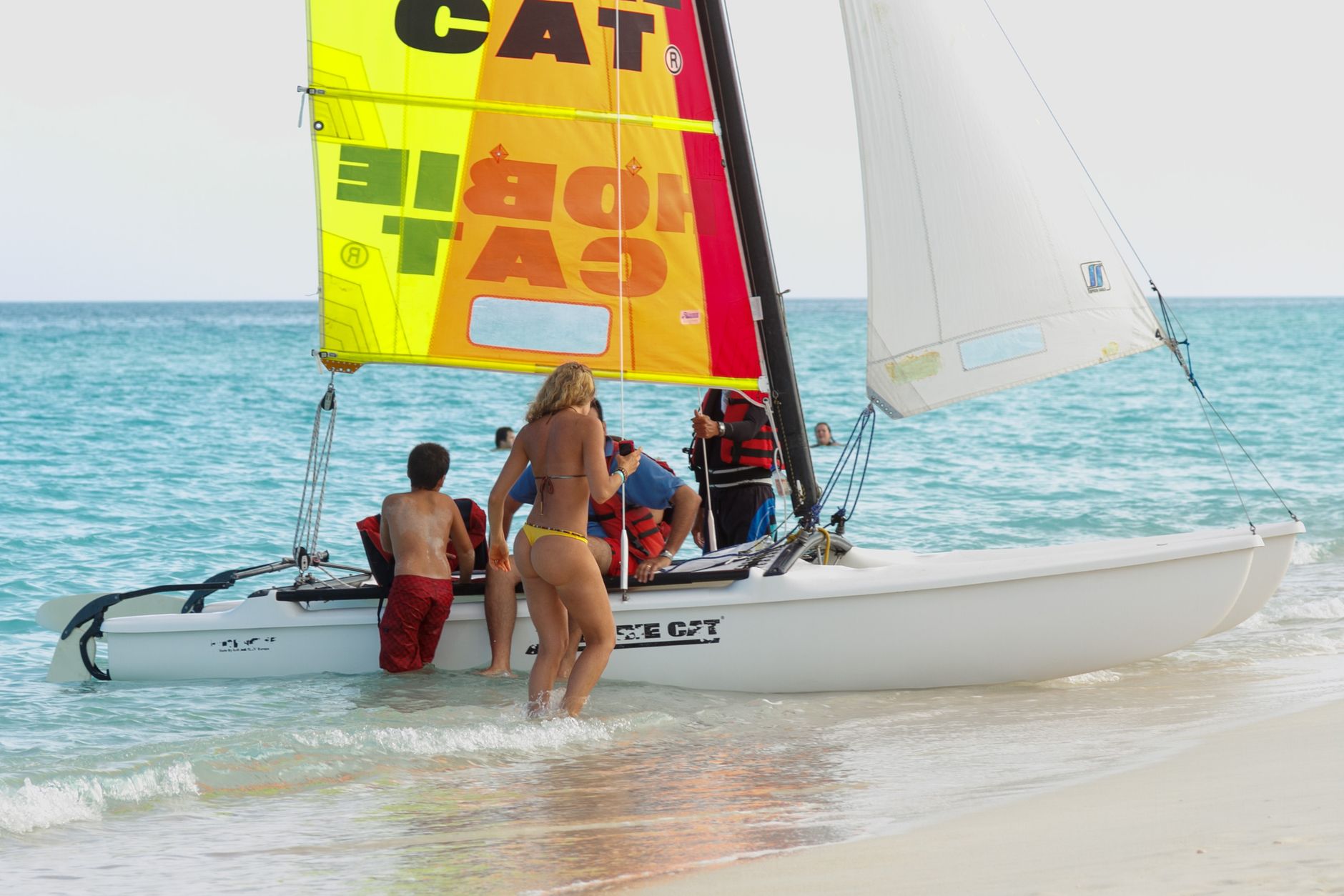 Young adults and children sailing a catamaran on a sunny beach day, enjoying summer fun.