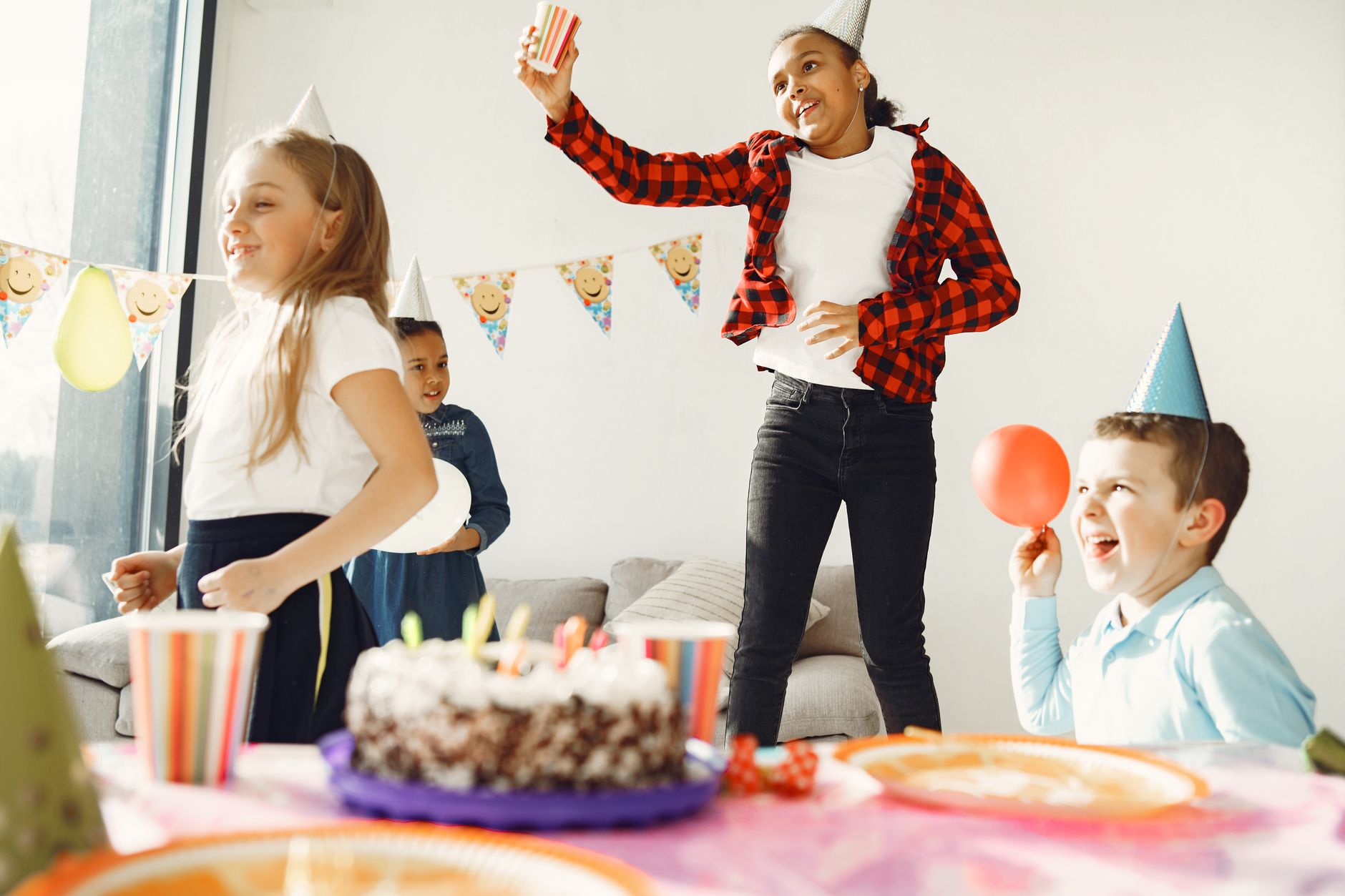 Children having fun at a birthday party with hats, cake, and balloons indoors.