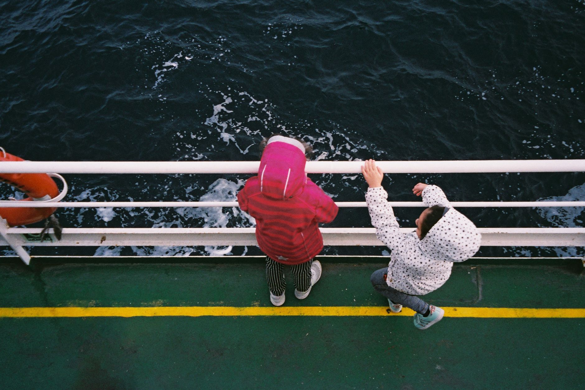 Two children in jackets stand on a boat deck, peering over the railing at the ocean.